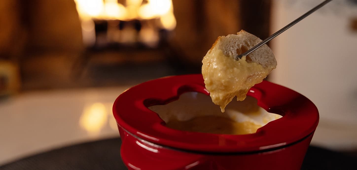 Bread dipped in cheese fondue, held over a red ceramic pot near a fireplace.