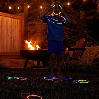 Child with glow stick necklaces and rings near a fire pit in a yard at night.