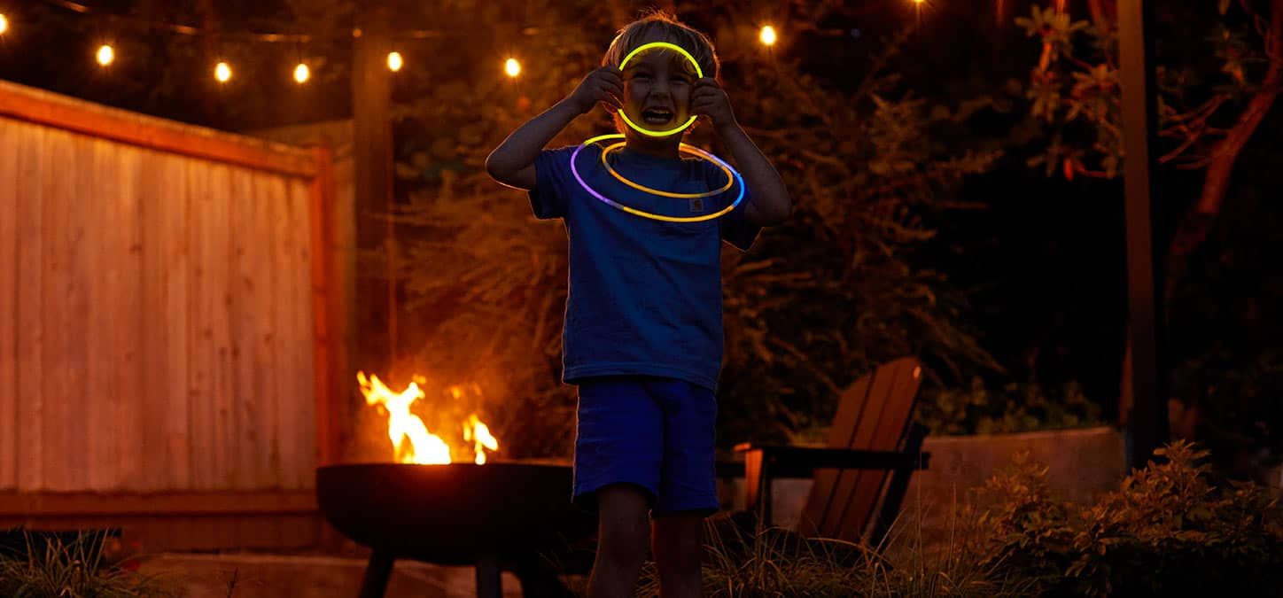 A young boy wearing glow in the dark necklaces is standing in front of a fire pit.