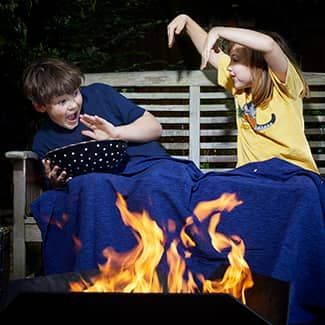Two children, blanket-covered, near a fire. One holds popcorn, the other gestures with excitement, telling a ghost story