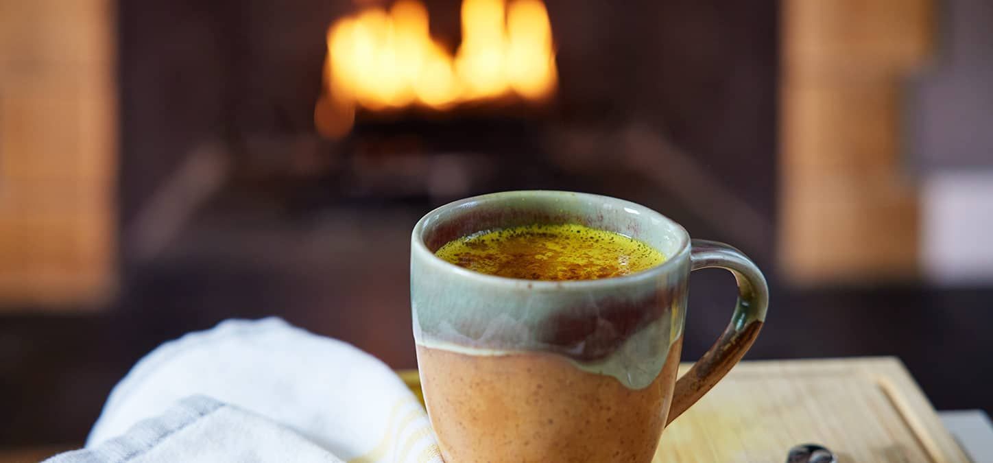 A mug of gold almond milk latte is sitting on a table in front of a fireplace.