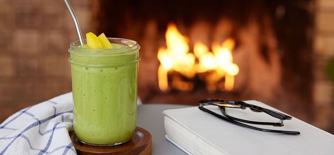 Green smoothie with mango garnish, near a fireplace, book, and glasses.