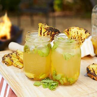 Two mason jars filled with pineapple mojito with grilled pineapple slices as a garnish, with a fire pit in the background