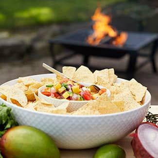 Chips with pineapple salsa in a bowl; mango, lime, and red onion on table; fire pit in background.