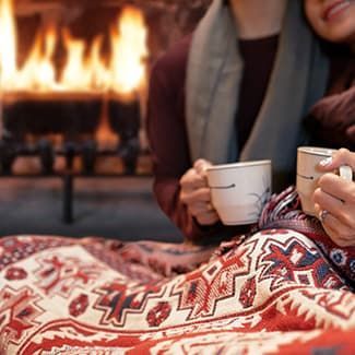 Couple by a fireplace, holding mugs, wrapped in a blanket with ornate red and white patterns.
