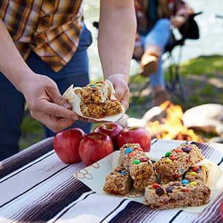 Plate of granola bars , apples, and bars on a picnic table. Fire and seated person in background.