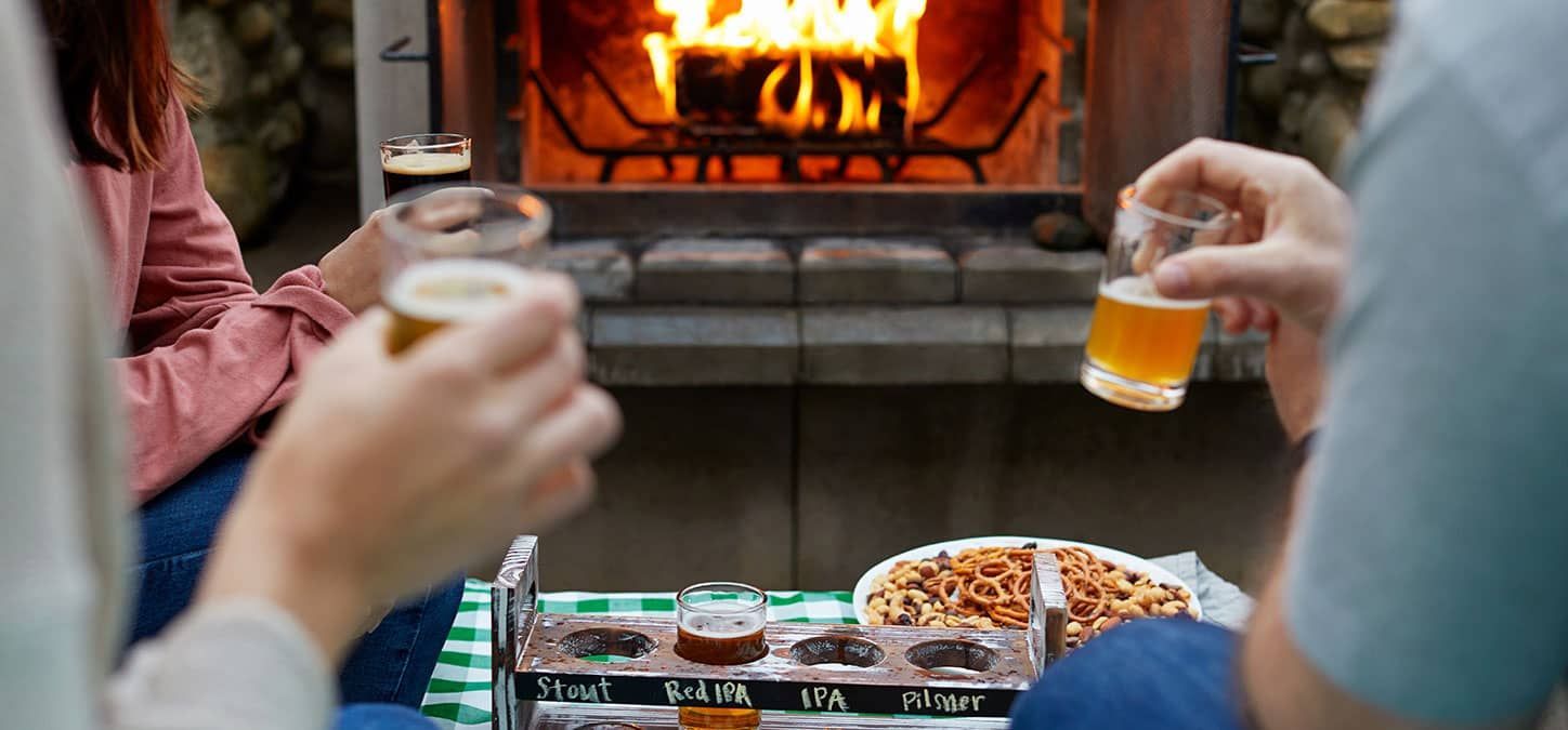 A man and a woman are toasting with beer in front of a fireplace.