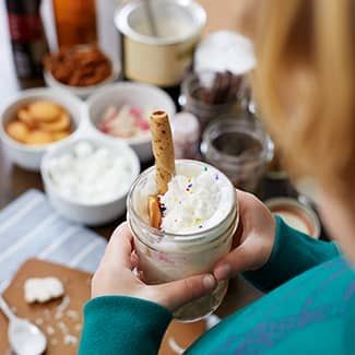 A person is holding a jar of cocoa with whipped cream and cookies in it.