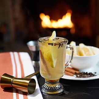 A hot toddy with a slice of lemon and cinnamon sticks on a table in front of a fireplace.