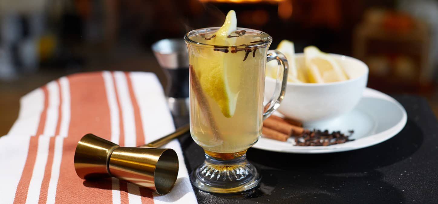 A hot toddy with a slice of lemon and cinnamon sticks on a table in front of a fireplace.