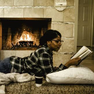 Person reading a book by a fireplace, lying on a blanket and pillow, wearing glasses and a plaid shirt.
