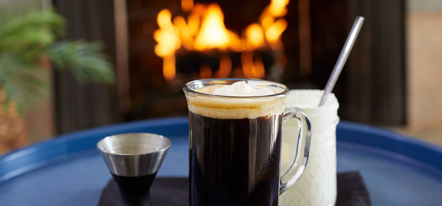 A cup of Irish coffee and a cup of whipped cream are on a table in front of a fireplace.