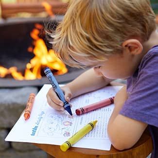 A young boy is sitting on a stool drawing on a piece of paper with crayons.