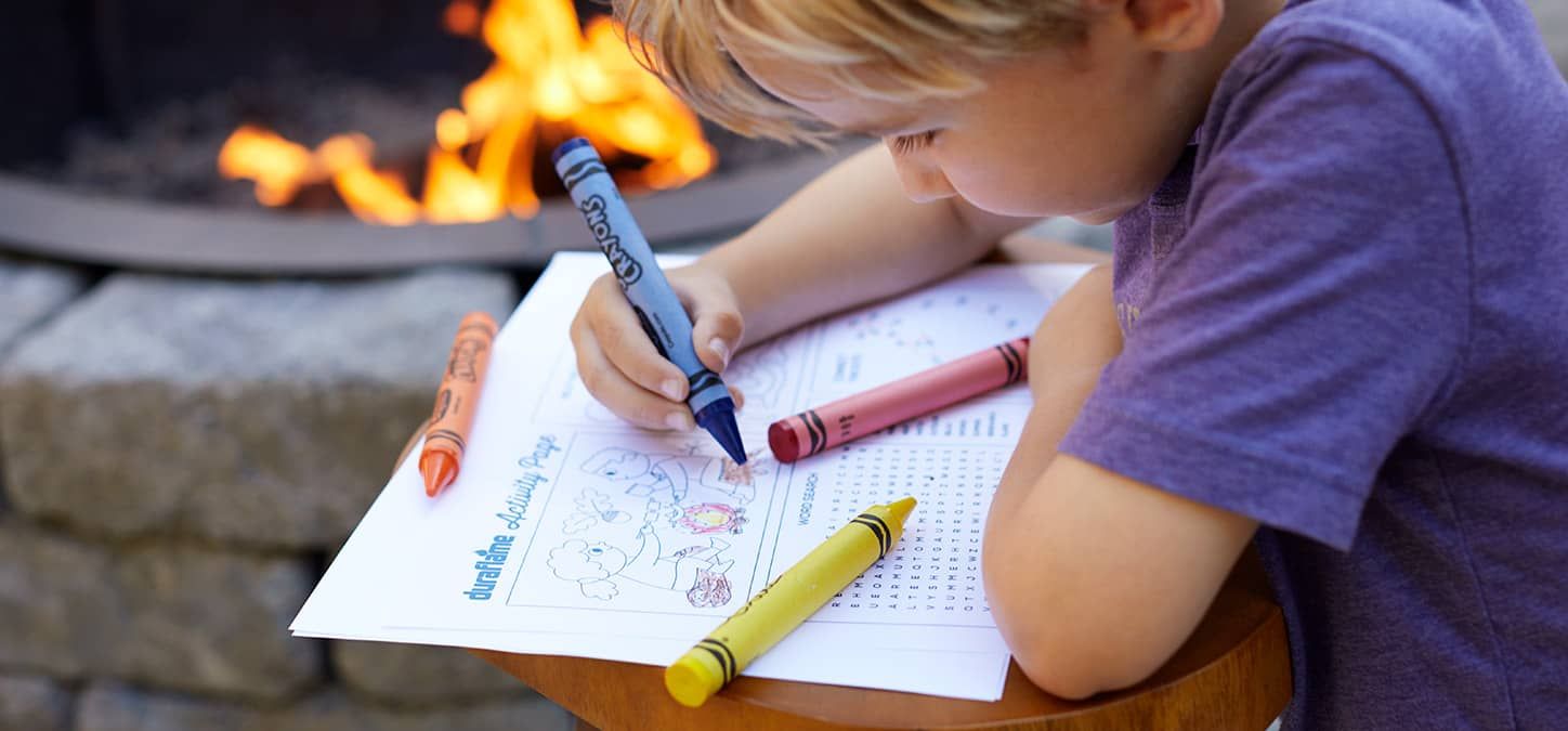 A young boy is sitting in front of a fire pit drawing with crayons.