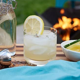A lavender gin sour with a slice of lemon on top of it is on a table in front of an outdoor fire