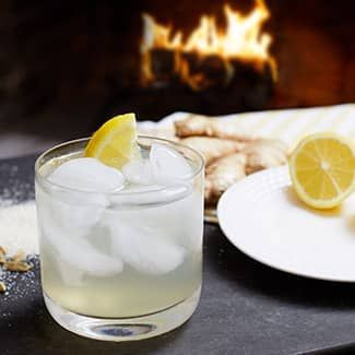 A glass of lemon ginger shrub mocktail with ice and a lemon slice on a table with a fire in the background