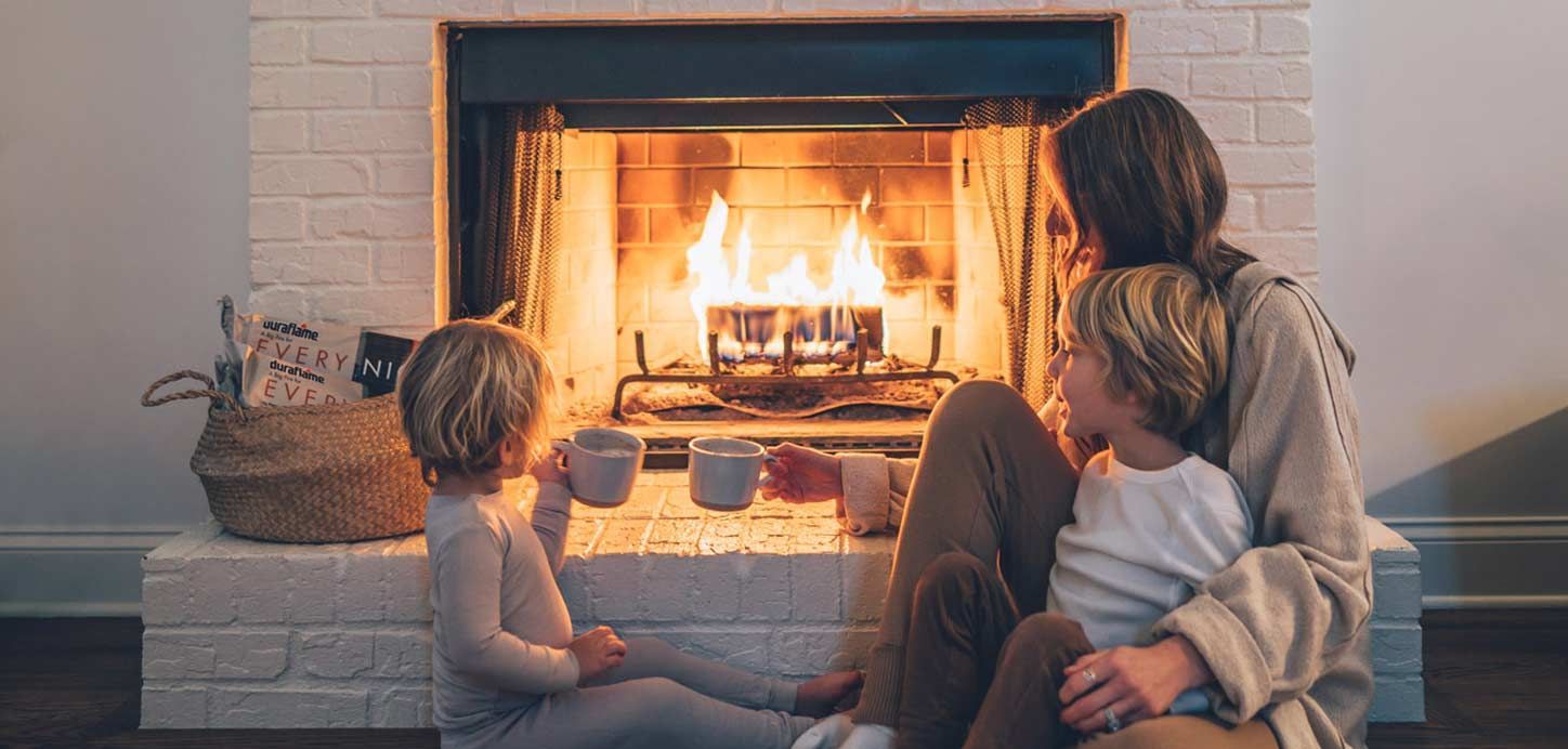 A mother and two children sit by a fireplace. One child drinks from a mug, as they watch the flames.