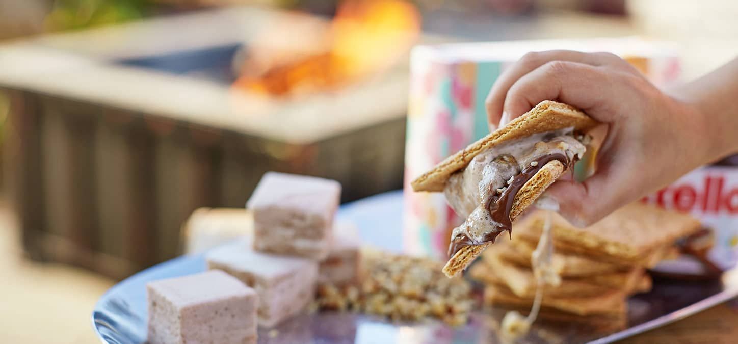 A person is holding a nutty about s'more with marshmallows and graham crackers in front of a fire pit