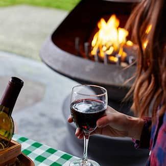 Woman holding wine glass near a fire pit with flames, bottle, and picnic table.