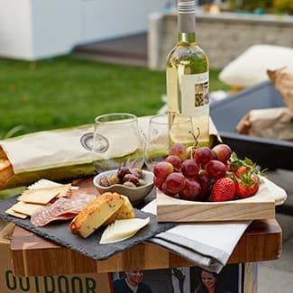 Picnic with wine, baguette, cheese, grapes, olives, and strawberries on a wooden tray outdoors.