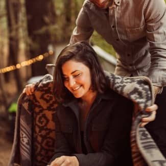 Man placing blanket on woman seated outside; smiling, warm tones.
