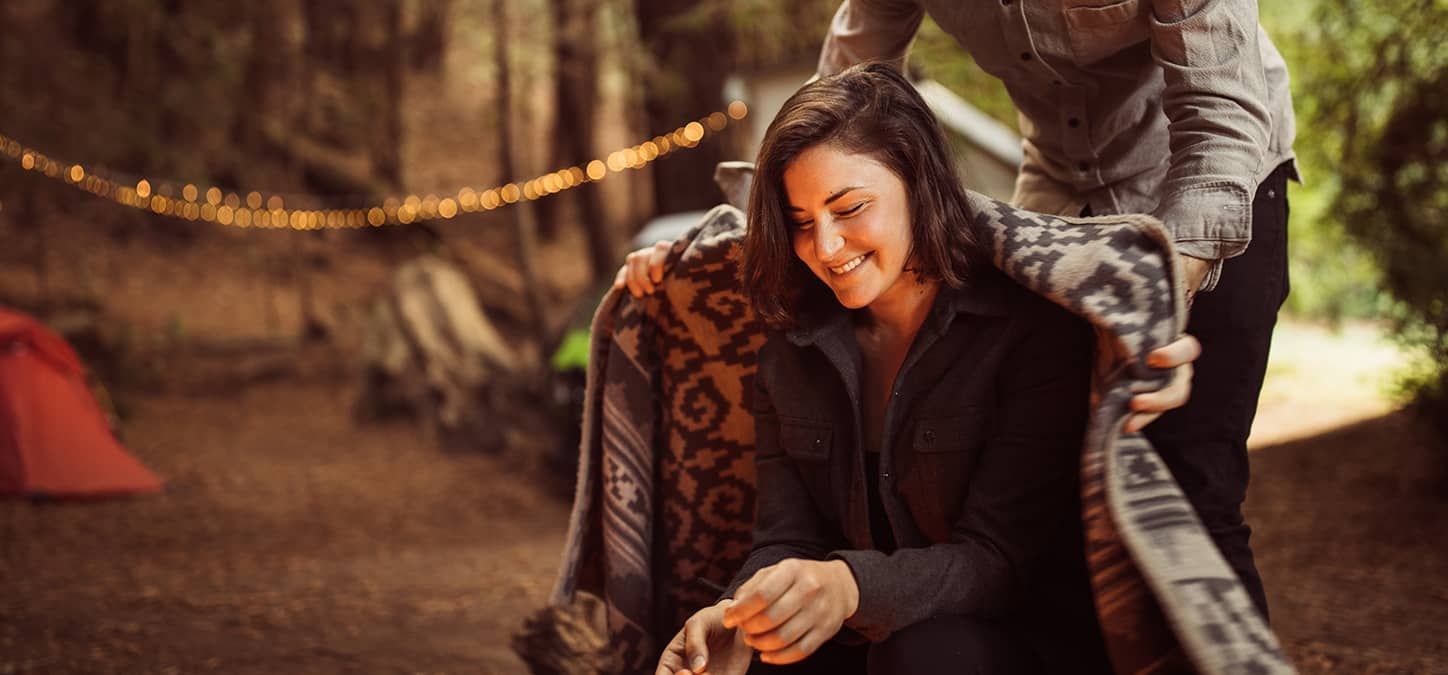 Man wrapping blanket around woman's shoulders fireside at campsite