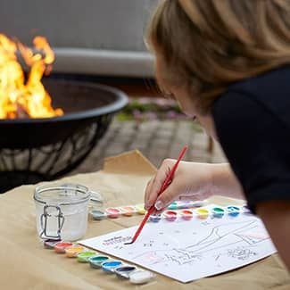 A child is painting a picture with watercolors on a piece of paper next to a fire pit