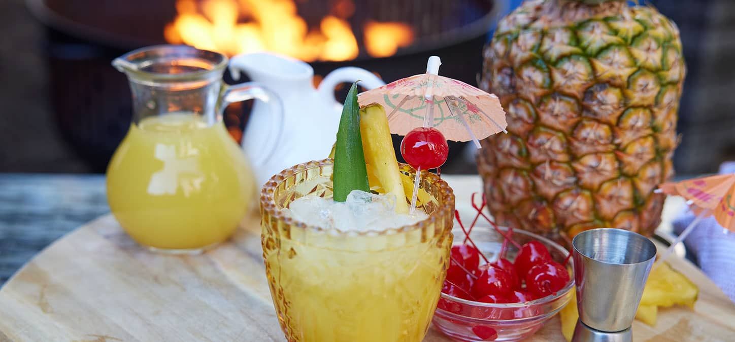 A pina colada cocktail with cherries and an umbrella on a wooden table in front of an outdoor fire