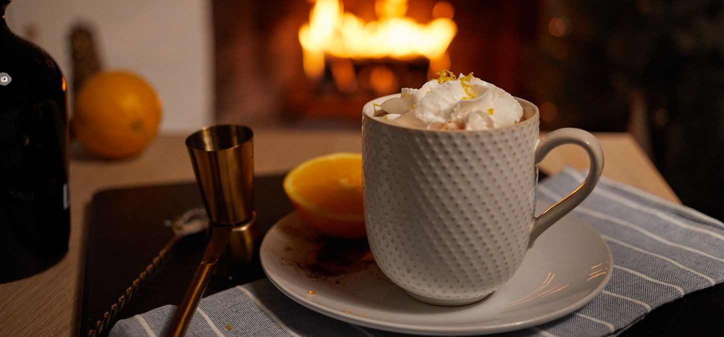 A cup of spiced orange hot chocolate with whipped cream is on a saucer in front of a fireplace.