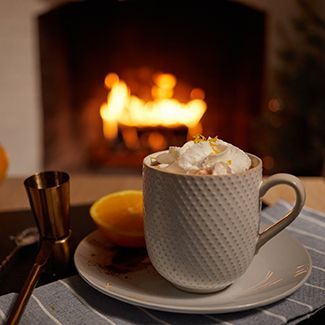 A mug of hot chocolate with whipped cream, orange slice, and a fireplace in the background.