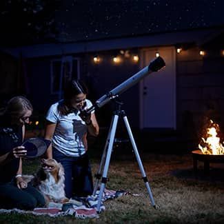 Two people and a dog stargazing with a telescope in a backyard at night, near a fire pit.