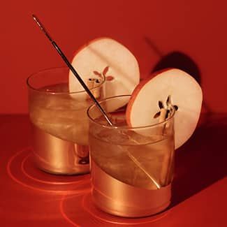 Two glasses of apple cider cocktail with straws and apple slices on a red table.