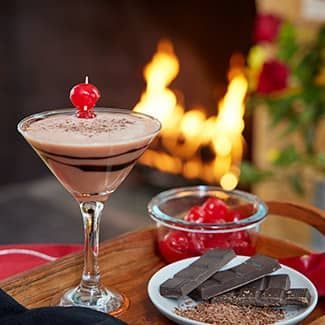 Chocolate martini with cherry garnish, chocolate bars, and cherries near a fireplace.