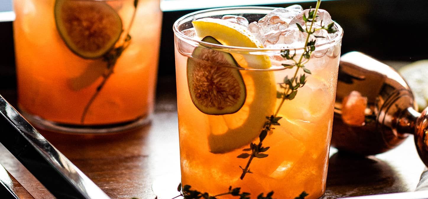 A close up of two spiced fig bourbon sours on a tray on a table in front of a fire