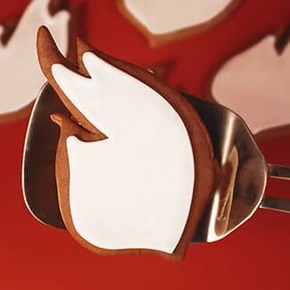 Dove-shaped cookie, brown outline, white frosting, held in a metal spatula against a red background.