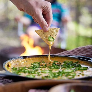 A person is dipping a tortilla chip into a skillet of cheese dip with a fire pit in the background
