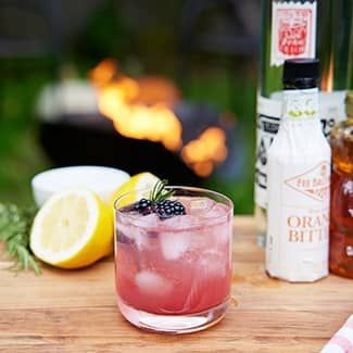 A glass of a spooky blackberry smash cocktail in a glass on a table in front of a fire pit