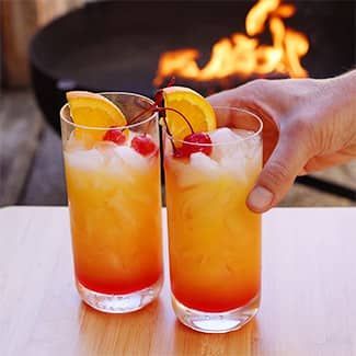 Two glasses of Tequila Sunrise with cherries and orange slices and a fire pit in the background