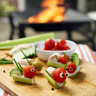 Celery snacks with tomatoes, olives, and chives (that look like friendly garden bugs) on a wooden board; grill in the background.