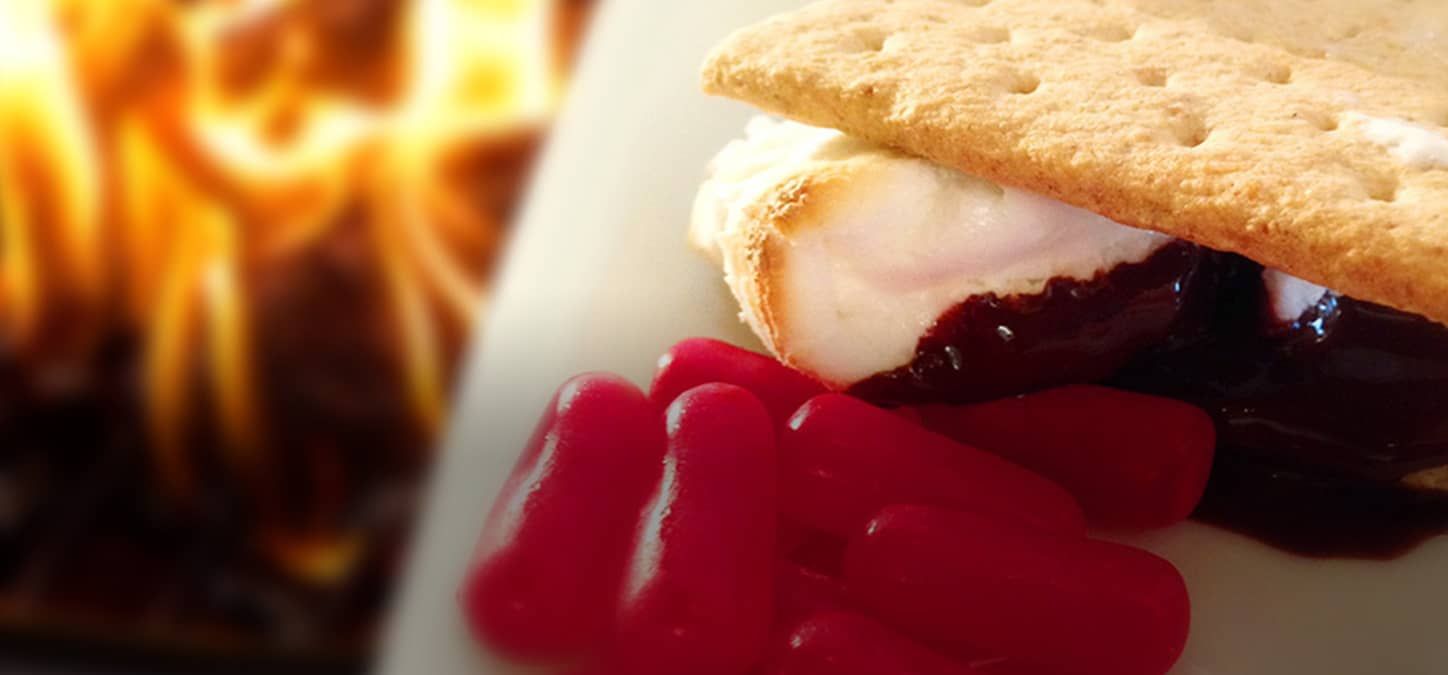 A s'mores sandwich with cinnamon candies on a plate with a fire in the background.