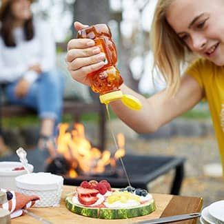Girl pouring honey from a bear-shaped bottle onto a watermelon pizza outdoors, near a fire.