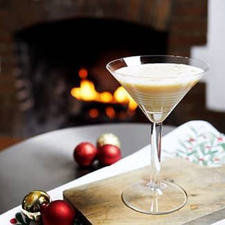 Martini glass with creamy drink on wooden board, Christmas decorations, fireplace in background.