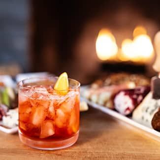 A negroni cocktail with a lemon wedge, ice, and charcuterie on a wooden surface with a fireplace in the background.