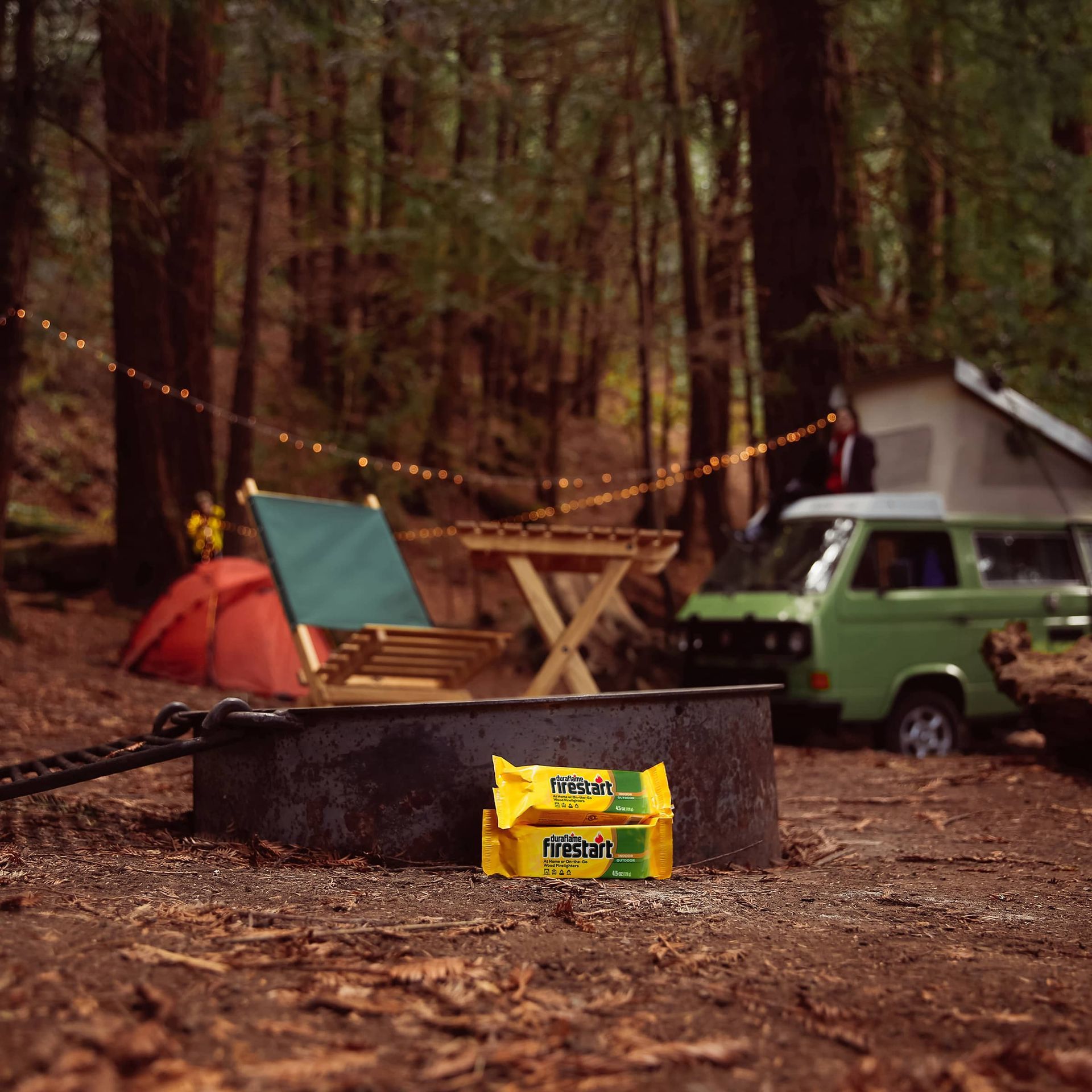 A green van is parked in the woods next to a fire pit with duraflame firestart firelighters in front