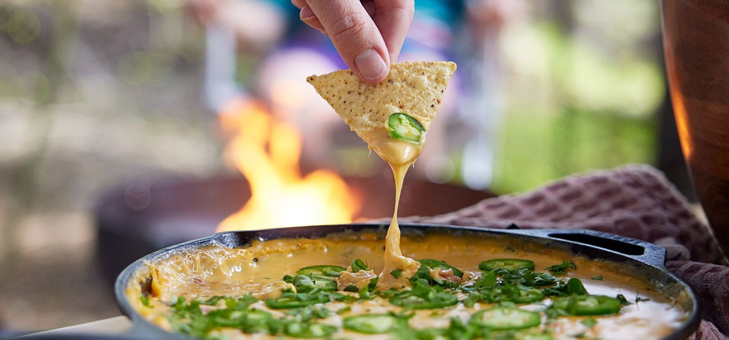 A person is dipping a tortilla chip into a bowl of dip in front of a fire pit