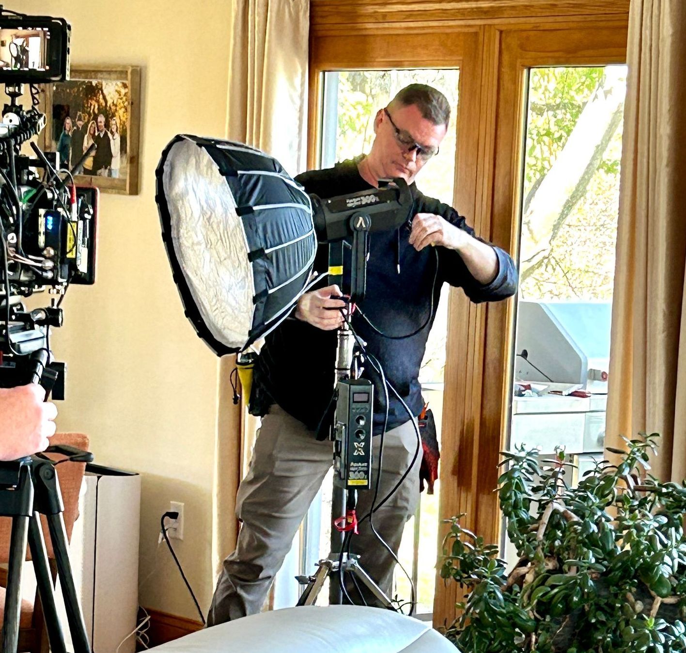 A person setting up a professional video lighting rig with a softbox in a room with a large window and houseplant.