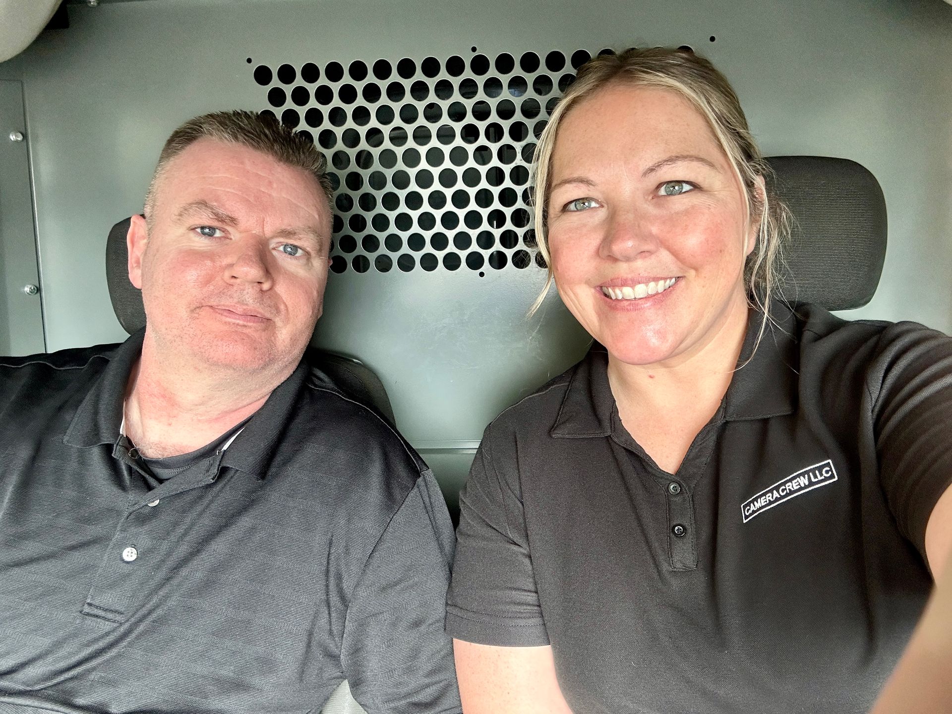 Two people wearing gray polo shirts pose for a selfie inside a vehicle with a metal partition behind them.