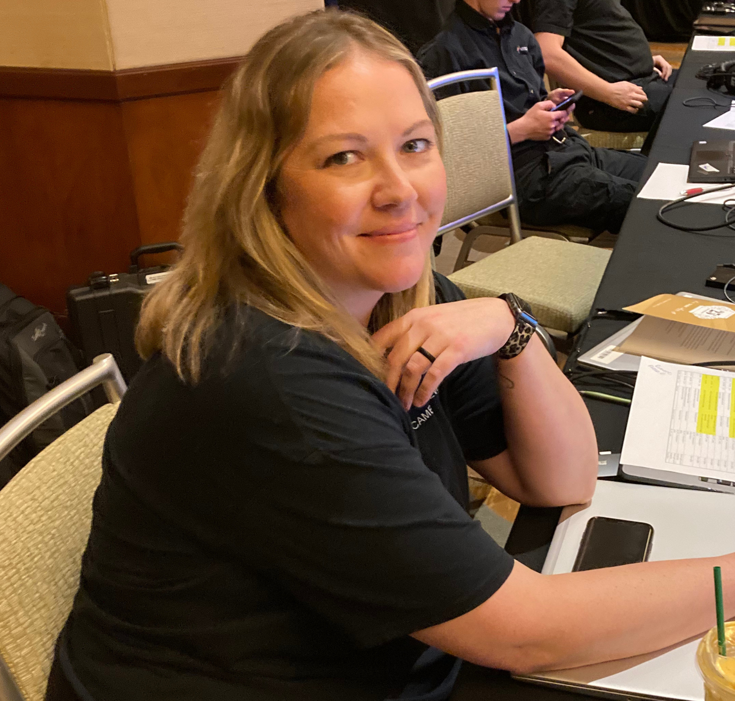 A person with shoulder-length blonde hair smiles while sitting at a table with a laptop, notebooks, and a drink.