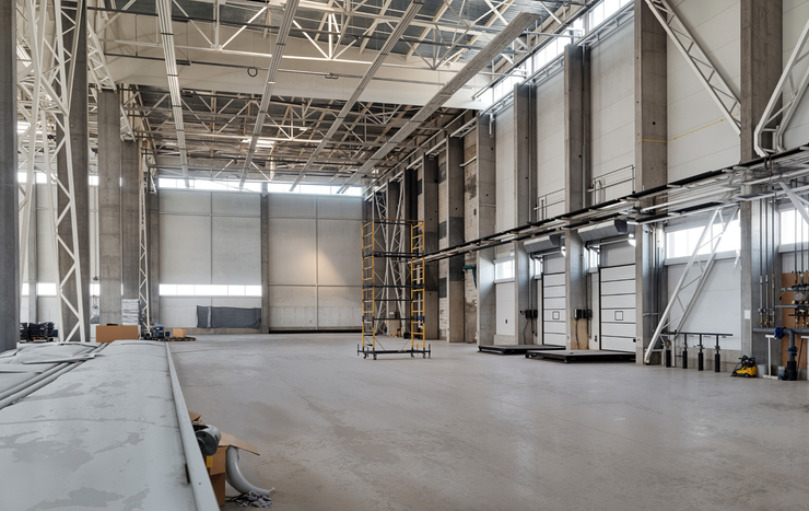 A vast, empty industrial warehouse with high ceilings, concrete pillars, exposed structural beams, and loading bays.