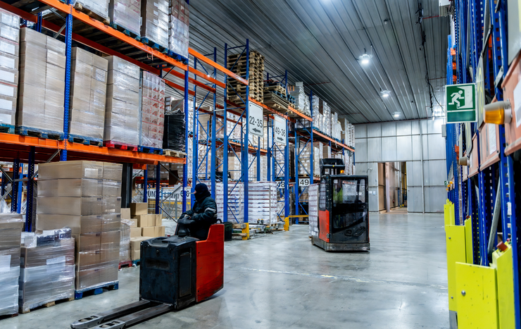 Warehouse interior with rows of tall storage racks filled with boxes and two forklift operators working.